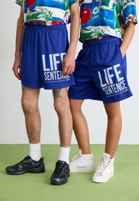Mėlynos tinklinės šortai su baltais užrašais "GYVENIMO BAUSMĖ". Du modeliai dėvi skirtingus batus, įskaitant juodus ir baltus sportbačius.