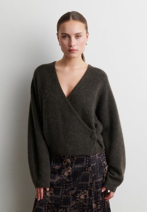 Vrouw met naar achteren getrokken haar draagt een donkergrijze, donzige wikkeltrui en een rok met patroon, staand tegen een effen lichte achtergrond.