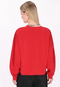 Felpa rossa, oversize, con scollatura arrotondata e maniche lunghe e larghe. Dotata di orlo a coste e realizzata in morbido cotone.