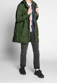 Grön lättviktsparka med huva, som har framfickor och en avslappnad passform, kombinerad med en randig skjorta och mörka jeans. Svarta sneakers.