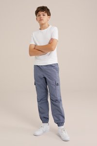 Bílý tričko s krátkým rukávem, šedé cargo kalhoty s elastickými manžetami, bočními kapsami a textilní etiketou. Bílé tenisky dotvářejí outfit.