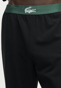 Gros plan sur un short noir avec une ceinture verte arborant un logo crocodile blanc au centre devant.