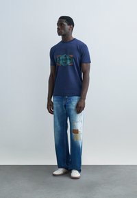 Άνδρας που φοράει μπλε ναυτικό t-shirt με γραφικά, ξεβαμμένα μπλε τζιν με μπεζ και καφέ μπαλώματα και λευκά αθλητικά παπούτσια, στέκεται μπροστά σε ένα απλό ανοιχτό γκρίζο φόντο.