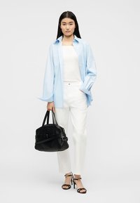 Femme debout portant une chemise bleu clair sur un haut blanc et un pantalon, tenant un sac à main noir, et portant des sandales à talons hauts noires.