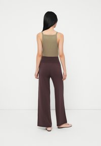Femme debout, de dos, portant un débardeur olive, un pantalon large taille haute marron et des chaussures plates rose clair, sur fond blanc uni.
