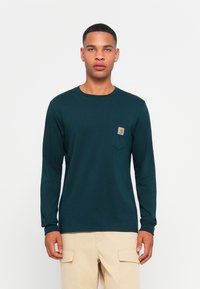 Lange mouwen, donkergroene shirt van katoen. Beschikt over een klein borstzakje met een logo en een ontspannen pasvorm. Gestructureerde stof met een gladde afwerking.