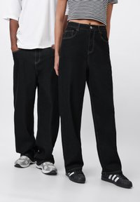 Svarte vide jeans med kontraststing og klassisk 5-lommers design, sammen med grå og svarte joggesko.
