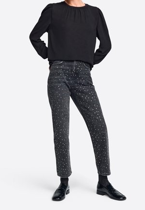 Person wearing black long-sleeve blouse, high-waisted black jeans with white speckled pattern, black socks, and black loafers, standing with hands behind.
