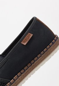 Chaussure noire en tissu à enfiler avec des accents en cuir marron et une semelle tressée. Présente des panneaux latéraux élastiques et un logo en relief sur la languette.