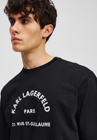 Juoda medvilnė ilgomis rankovėmis marškinėliai su baltais kreivais užrašais: "Karl Lagerfeld Paris 21 Rue St-Guillaume." Minkšta tekstūra, apvalaus kaklo dizainas.