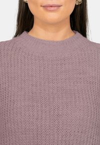 Vrouw met gouden oorringen en een lichtpaarse grofgebreide trui met geribde kraag, getoond van kin tot schouders.