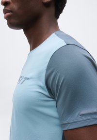 Άνδρας που φοράει ανοιχτό γαλάζιο αθλητικό μπλουζάκι με πιο σκούρα μπλε μανίκια, εμφανιζόμενος από το λαιμό μέχρι τη μέση του κορμού με φόντο μονόχρωμο.