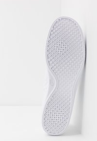 Pantofă sport albă cu o talpă din cauciuc texturat, având gripuri circulare. Pe lateral se află un logo subtil. Designul superior este dintr-un textil neted.