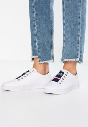 Witte slip-on sneakers met marineblauwe, rode en witte gestreepte elastische banden, gedragen met lichtblauwe jeans met rafelige zoom op een witte vloer.