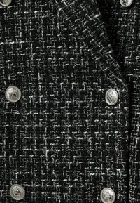 Melnā un baltā austā auduma ar teksturētu rakstu un metāliskiem pogu akcentiem, veidojot strukturētu dizainu.