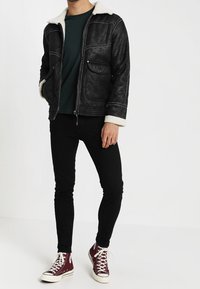 Svart shearling-fodrad jacka med två framfickor, bärs över en grön topp, kombinerad med svarta jeans och bordeauxfärgade sneakers.