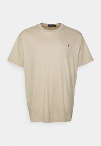 Beige tričko s krátkým rukávem vystavené na bílém pozadí, s malým logem na levé části hrudi.