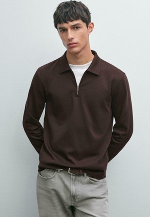 Jonge man in een donkerbruine ritsloze lange mouw polo over een wit shirt en lichtgrijze broek, staande met de handen achter zijn rug.