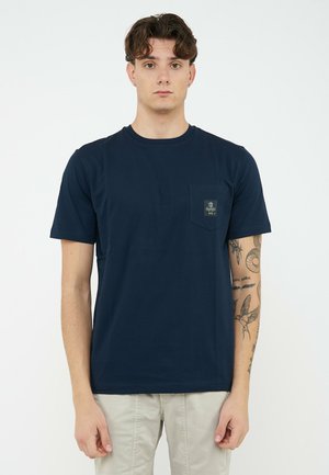 Giovane uomo con capelli corti e ricci che indossa una maglietta blu navy con tasca e pantaloni beige su uno sfondo bianco uniforme.