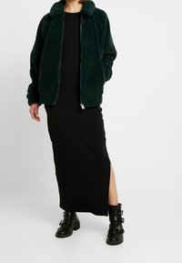 Zielona polarowa kurtka bombowiec z zamkiem i kołnierzem, noszona na czarnej sukience midi z bocznym rozcięciem. Czarne masywne botki dopełniają outfit.