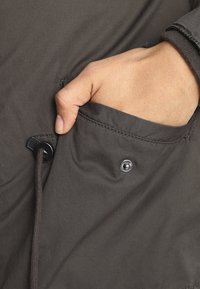 Ciemnoszara tkanina kurtki z widoczną ręką w przedniej kieszeni. Posiada sznurki i zapięcie na zatrzask w kieszeni. Gładka tekstura.