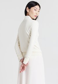 Cremfarbener Pullover mit rundem Ausschnitt und geripptem Saum, kombiniert mit einem hellen Rock. Strickstruktur, taillierte Passform und lange Ärmel.