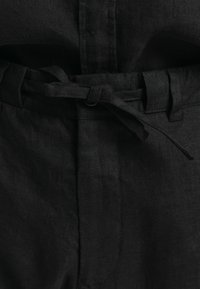 Svart linnackoverall med knytdetalj i midjan. Har en mjuk textur och en ren silhuett som framhäver en enkel och funktionell design.