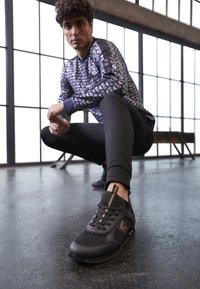 Man som hukar inomhus, iförd svarta och guld EA7-sneakers, svartabyxor och en långärmad skjorta med blå EA7-logotypmönster.