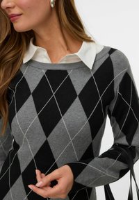Donna che indossa un maglione argyle grigio e nero sopra una camicia bianca con colletto, con orecchini ovali d'argento e che regge il manico di una borsa nera.