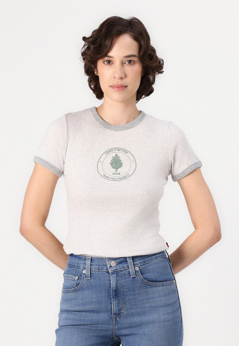 Frau mit kurzen, lockigen Haaren, die ein graues Ringer-T-Shirt mit einem grünen Baum-Logo und dem Schriftzug "Hinterlasse es besser, als du es vorgefunden hast" sowie blaue Jeans trägt.