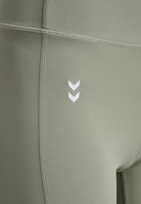 Olivgrüne Sportleggings aus glattem Material mit kontrastierenden weißen Chevron-Details und sichtbaren Flachnähten. Nahaufnahme.