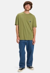 Olive green t-shirt with a chest pocket, textured cotton fabric, and a logo patch. Paired with blue loose-fitting jeans and black sneakers.