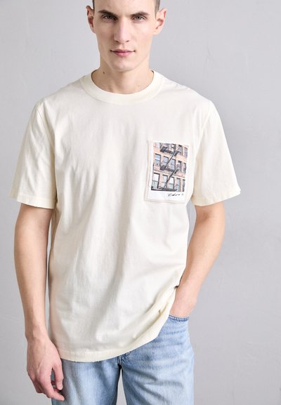 Kreemi värvi puuvillane t-särk, millel on ees tasku, millel on graafiline joonistus hoonest. Lühikesed varrukad ja lõdva istuvuse disain.