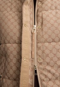 Beige wattierte Jacke aus strukturiertem, gemustertem Stoff und einem auffälligen goldenen Reißverschluss mit Logo-Akzent entlang der Verschlusskante.