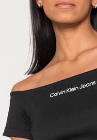 Kvinna med svart off-shoulder topp med vit text "Calvin Klein Jeans", silverfärgade hoopörhängen och rakt svart hår.