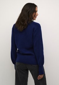 Pulover tricotat de culoare albastru marin cu o finisare texturată, margine și manșete cu dantele și o croială lejeră. Purtați-l cu pantaloni gri închis.