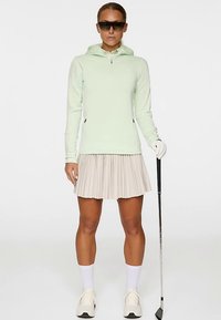 Felpa con zip verde chiaro, gonna beige a pieghe, calzini bianchi alla caviglia, guanto da golf e mazza. Realizzato per abbigliamento sportivo con un'estetica moderna.