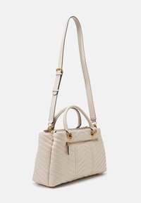 Sac à main beige matelassé avec un motif en chevrons, poignées supérieures, une bandoulière amovible, des accessoires en métal doré et une poche zippée à l'avant.