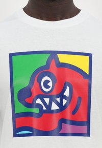 Weißes T-Shirt mit einem lebendigen roten Kopf eines Wesens, umrandet von marineblauen Linien, umgeben von grünen und gelben Blöcken, mit scharfen weißen Zähnen.
