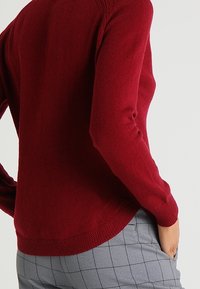 Czerwony sweter dzianinowy z długimi rękawami, zaokrąglonym dołem oraz teksturowanymi prążkowanymi akcentami na mankietach i na dole. Noszony z szarymi spodniami w kratę.