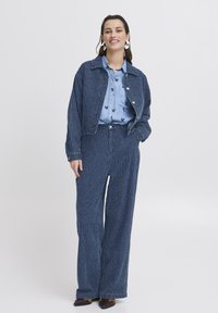 Mujer de pie vestida con una chaqueta azul a rayas y pantalones anchos a juego, con una camisa floral de un azul claro y grandes pendientes de aro.