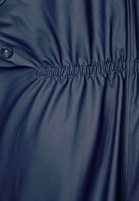 Tela de chaqueta impermeable de color azul marino con una textura suave, que presenta un elástico recogido en la cintura y un detalle de cierre a presión.