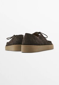 Chaussures en daim marron foncé avec bout rond, lacets à l'avant et semelle en caoutchouc beige. Caractéristiques : texture lisse et design minimaliste.