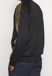 Svart sweatshirt med gyllene ränder längs ärmarna, dragkedjefickor på sidorna och ribbad nederkant och ärmslut; slät textur och atletisk design.