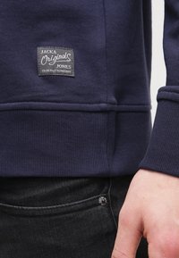 Manica e orlo di una felpa navy con patch del logo "Jack & Jones Originals" sopra la tasca dei jeans neri, con parte di una mano visibile.