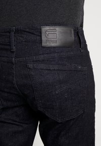 Jeans de un azul oscuro con un diseño ajustado, una textura sutil y una etiqueta de cuero negro con la marca "RAW" en la cinturilla.