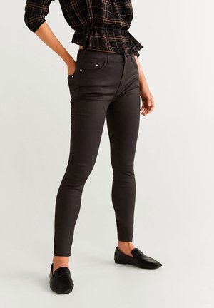 Frau trägt schwarze Skinny-Hose, schwarze Slipper und ein schwarzes kariertes Peplum-Top mit kurzen Ärmeln.
