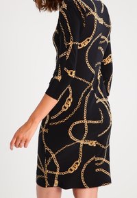 Sort kjole med trekvartærmer, pyntet med et guld kædemønster. Strækbart materiale, tætsiddende design og knælængde.