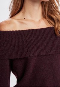 Žena v tmavě fialovém svetru bez ramínek s ribkovým vzorem, ukazující klíční kosti a část dlouhých hnědých vlasů.