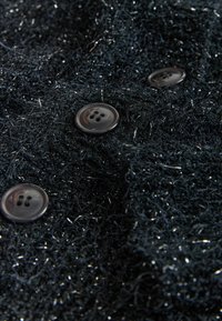 Black textured fabric with metallic threads. Features three glossy black buttons with four holes. Close-up view, highlighting material detail.
